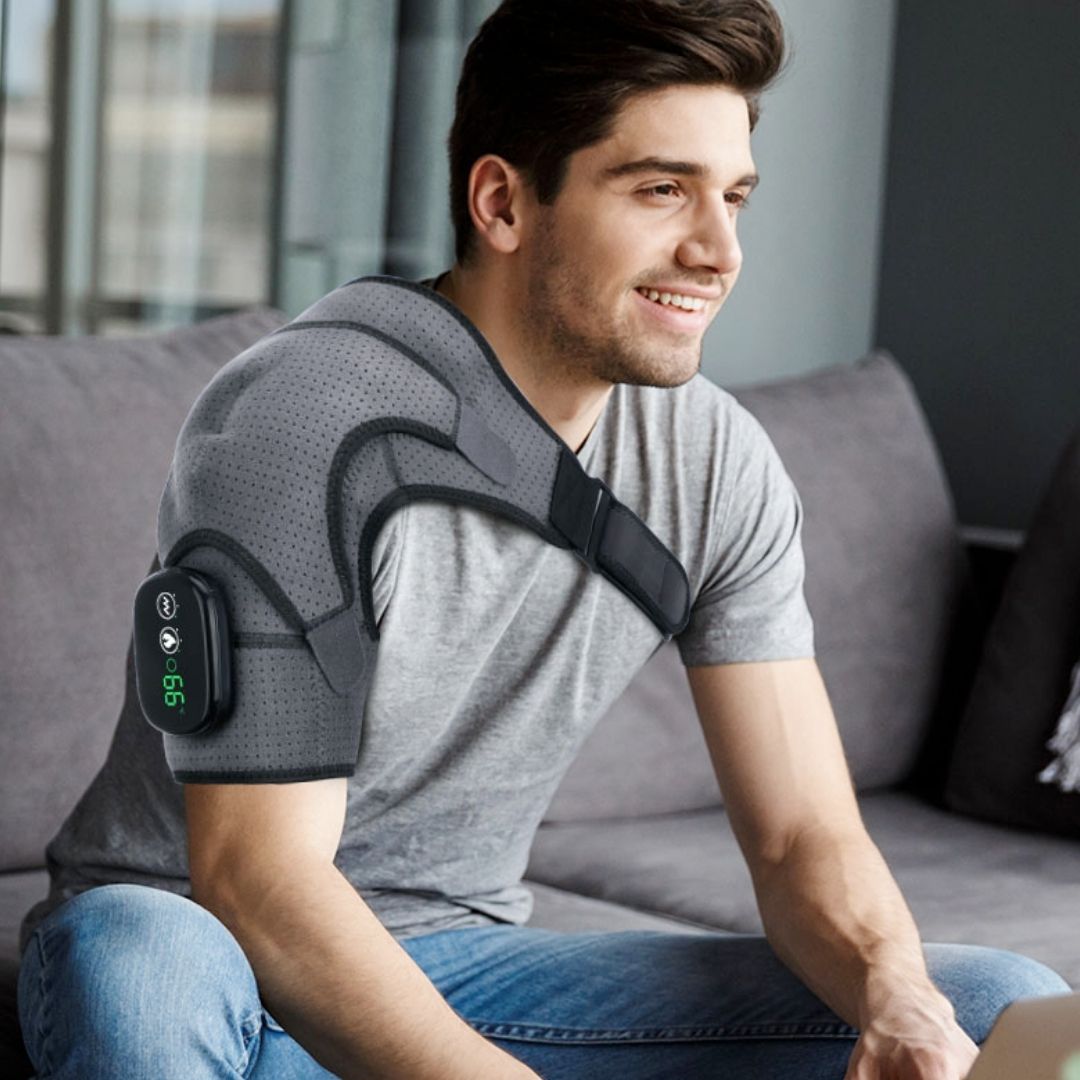 Smiling young man seated on grey sofa wearing heated compression shoulder brace with LED display