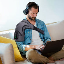 Load image into Gallery viewer, Man with headphones using laptop wearing grey heated compression shoulder brace on sofa