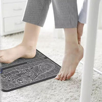 Person in striped trousers using EMS Foot Massage Mat on white carpet indoors