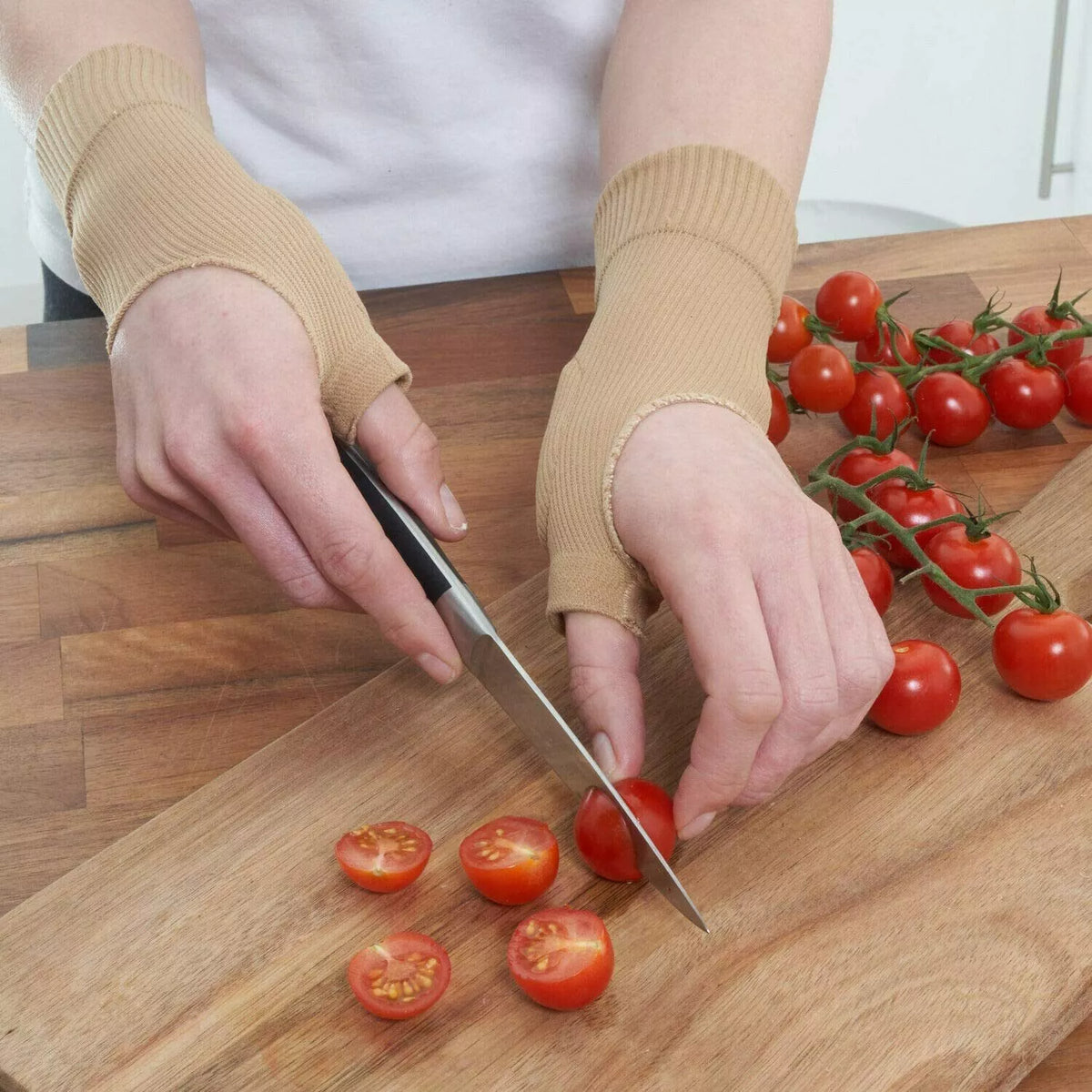 Person wearing beige wrist support gloves chopping cherry tomatoes on a wooden board