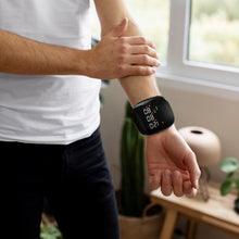 Load image into Gallery viewer, Man in white shirt checking black wrist blood pressure monitor indoors near a window and houseplants
