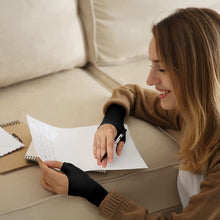 Load image into Gallery viewer, Smiling woman wearing black gel thumb support gloves writing a letter on a sofa
